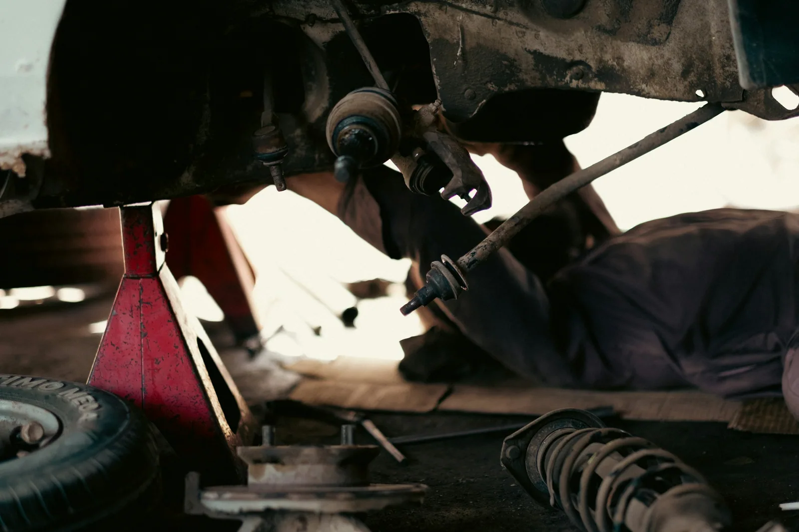 Arbeit am Fahrwerk: ein Mechaniker liegt unter einem aufgebockten Auto, davor Stoßdämpfer und Reifen