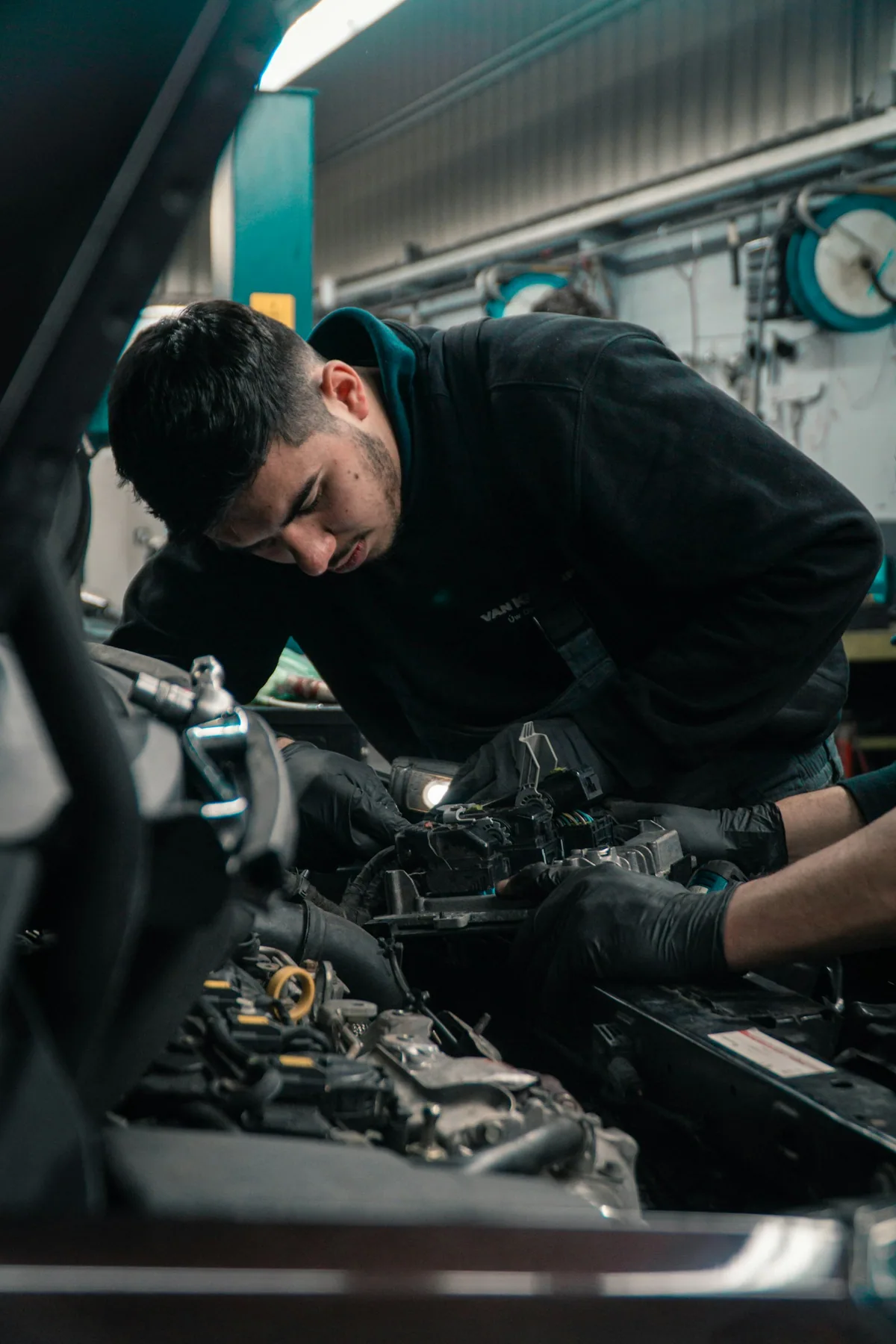 Ein Mechaniker beugt sich über den offenen Motorraum und arbeitet mit Handschuhen an der Elektrik