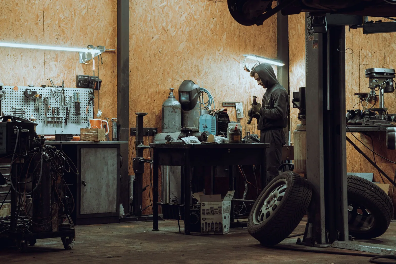 Werkstatthalle am Abend: ein Mechaniker an der Werkbank, davor Hebebühne und Reifen